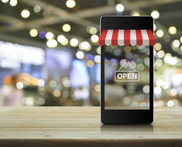 Modern smart mobile phone with on line shopping store graphic and open sign on wooden table over blur light and shadow of mall